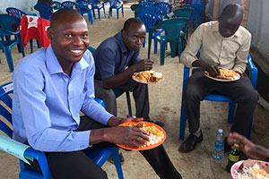 男性の先生たち3人が食事を食べながら笑顔でこちらを向いている