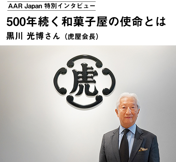 黒川会長が朗らかな笑みを浮かべている　後方の壁に大きな企業ロゴが見える