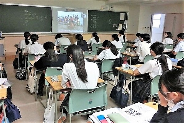 教室の中で生徒たちが椅子に座り、黒板にかけられたスクリーンに映るオンライン授業を見ている