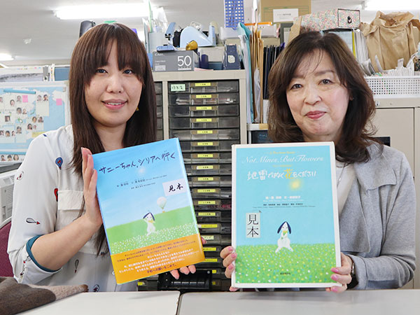 ボランティアの小柳澄枝さんとAR東京事務局の鎌田舞衣が、絵本地雷ではなく花をくださいシリーズを1冊ずつ持ち、笑顔で並んでいる