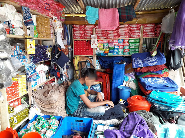 男の子がトタン屋根の小さなお店の中に胡坐をかいて座っている。周りには棚があり、UNHCRと書かれた復路などが所狭しと積まれている