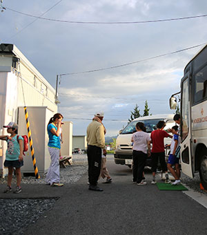 左に仮設住宅、右に大型のバス、その間に子どもたち数人や鎌田が写っている