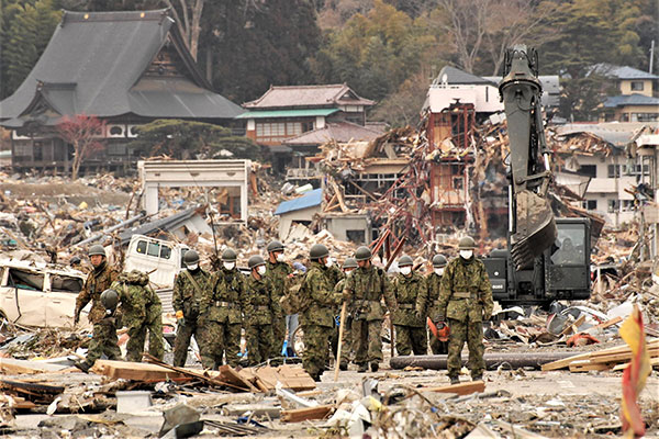 津波や地震の被害で瓦礫だらけの市街地を15名ほどの迷彩服を着てマスクをつけた自衛隊員が歩いている