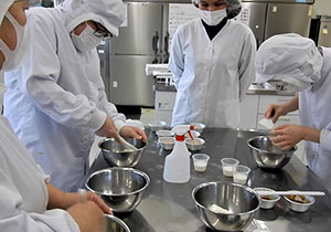 4人ほどの白い作業服を着た利用者が、ボールやハンドミキサーを手に取りお菓子作りをしている