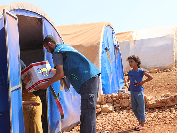難民キャンプで男性が食糧の入った段ボールをキャンプ内の女性に手渡している。女の子がその様子を右横でみている