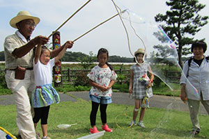 女の子シャボン玉作りにチャレンジ　松川浦サポートセンターのボランティアの男性が一緒にシャボン玉作りの棒を持ってサポート　女の子より大きなシャボン玉がうっすらとできている