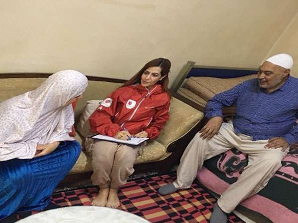 難民世帯を訪問し、家の中でAAR女性スタッフがノートとペンを持ち、聞き取りを行っている。左右には難民の女性と男性がソファに座り、質問に答えている