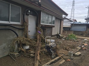 玄関前に泥まみれのシルバーカーや壁に流木、草などが流れ着いている住宅