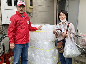 AAR緊急支援チームの大原と耕野地区世話人の刈田さんが、積まれた支援物資のポリタンクの前に並ぶ。刈田さんは支援物資の即席の味噌汁が入った袋を持つ