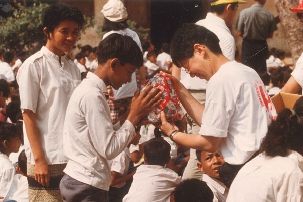 カンボジアでポシェットを手渡す男性と受け取る少年、後ろには大勢の子どもたち