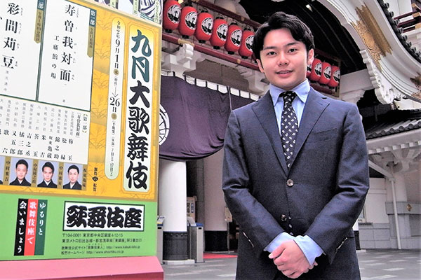 中村莟玉さんが歌舞伎座を背にして笑顔でこちらをみている　グレイのスーツ姿