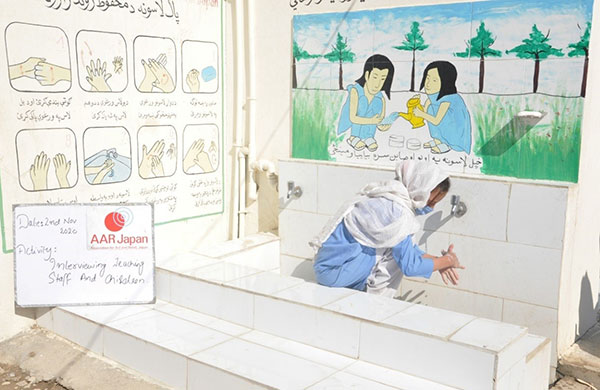 屋外の手洗い場で女の子が手を洗っている　壁面には、大きく手洗いの手順が描かれている