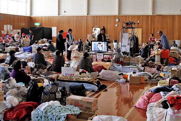 個々の布団、衣類、段ボールなどの荷物が雑然と広がっている避難所　中央にはテレビが設置されている