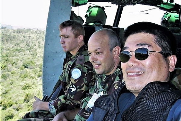 ヘリコプターの中に男性が3名座っており、外には空と草原が見える