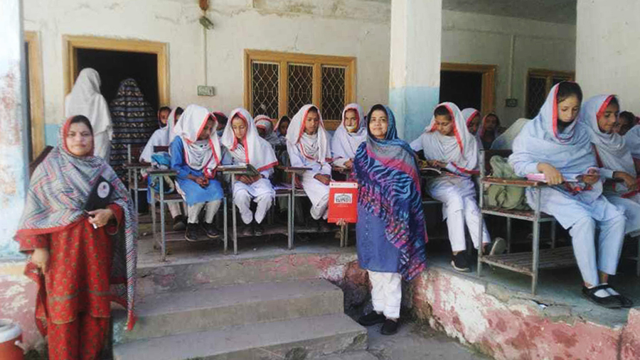 Many female students are sitting outside the classroom. Two teachers are standing beside them.