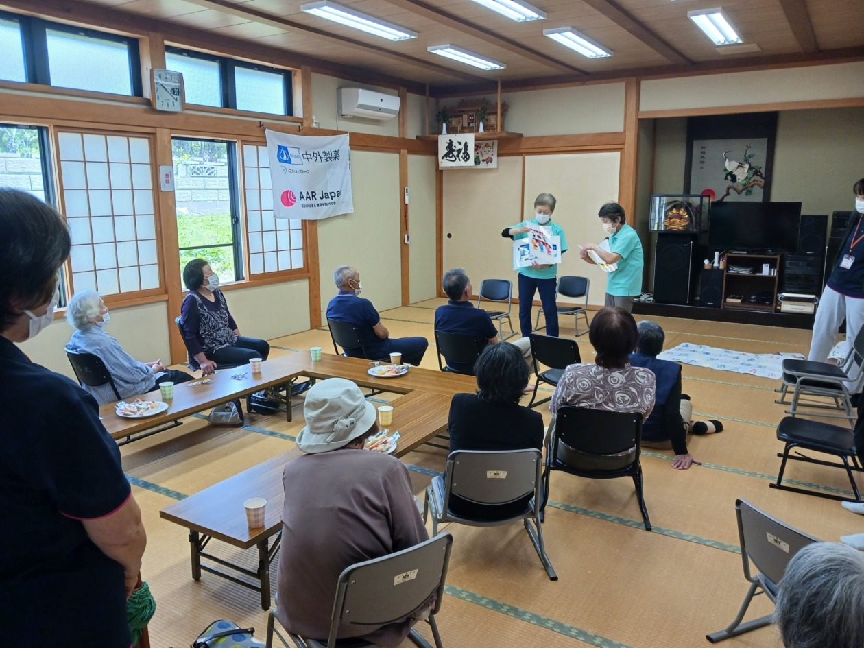 People sit in a community hall while two women give a presentation using illustrated materials