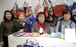 手芸品や和裁用品を前に４人の女性が出店しているところ。後方には色とりどりのカバンが陳列