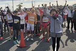 マラソン会場で手を高く上にあげてポーズをとる谷川さんと後方で同様にポーズをとるランナーの方々