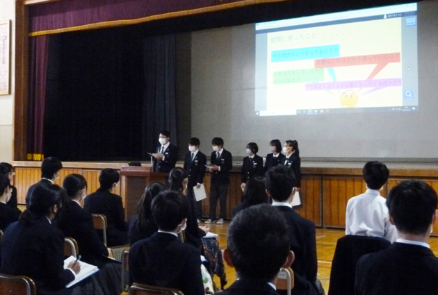 6名の生徒が体育館前方でスライドを投影しながら発表している