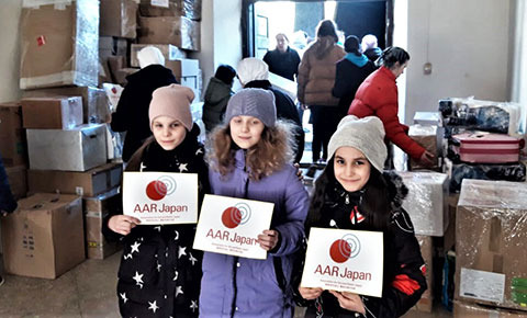 支援物資の前でAARのロゴを持ち笑顔の少女3人