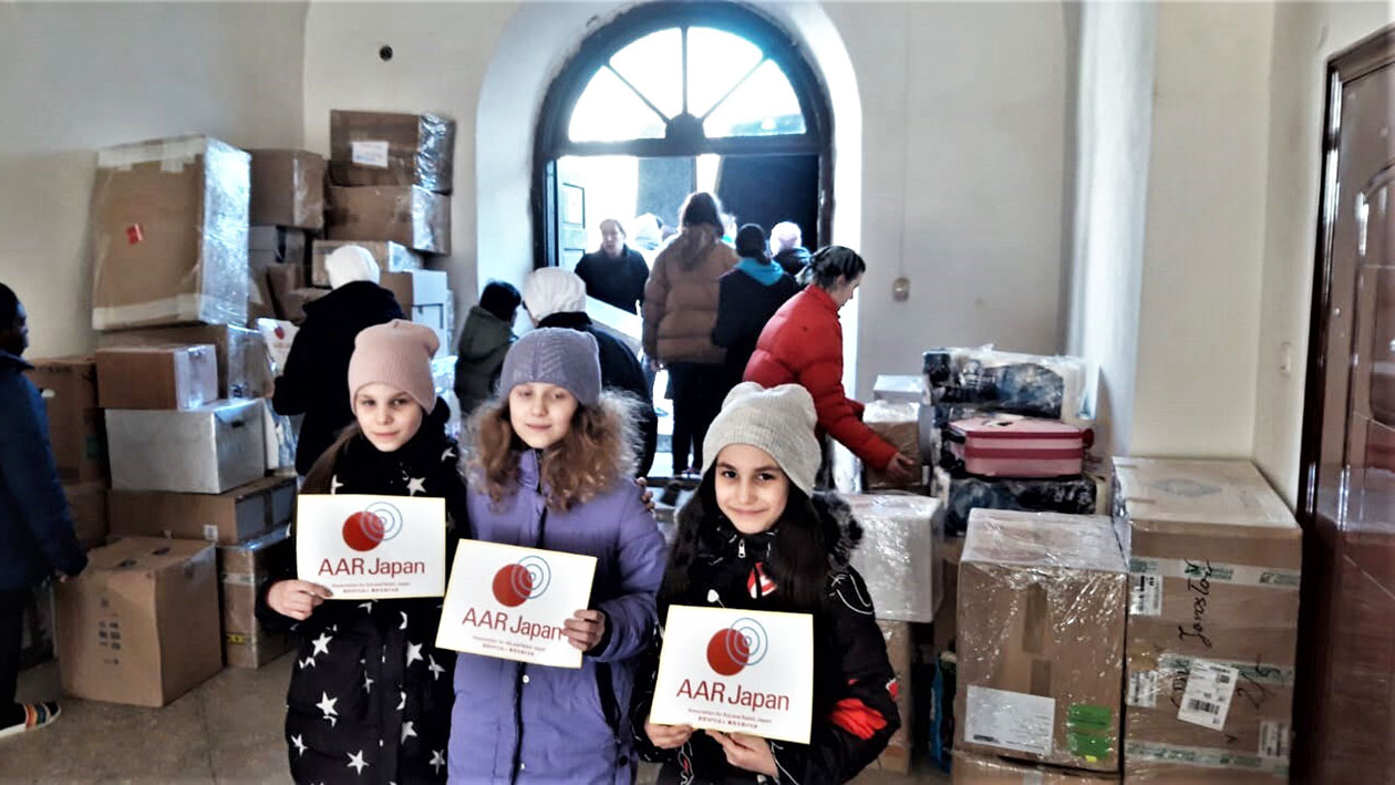 支援物資の横でAARのロゴを手にする少女3人　奥は物資を修道院に運び込む人たち