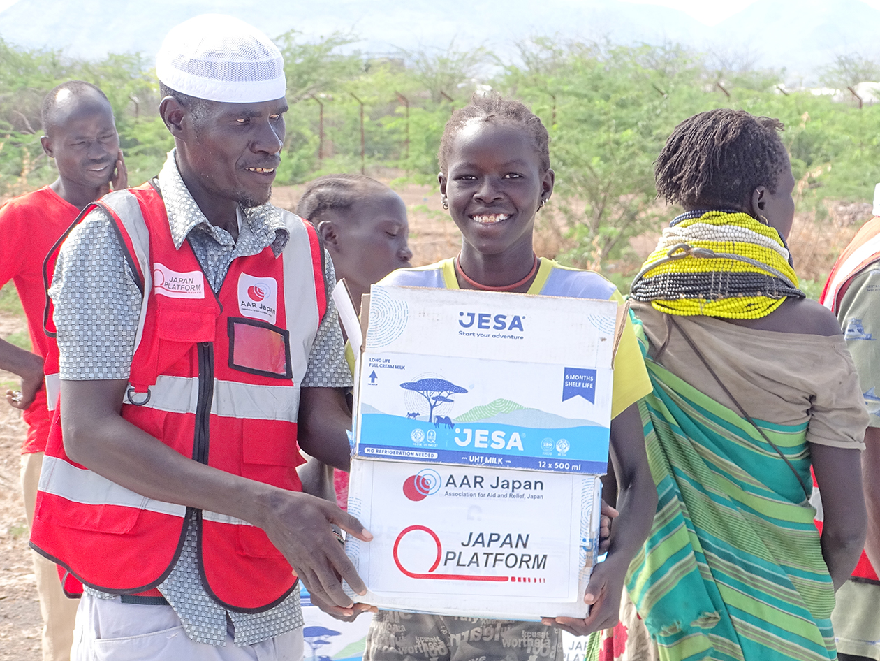 ケニアの女性が支援物資の入った箱2つを持って笑顔で立っている
