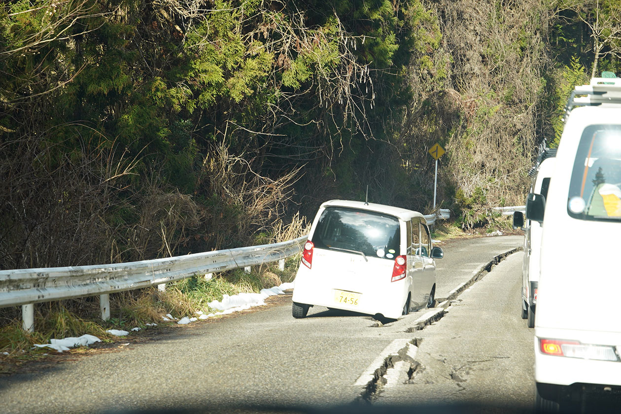 道路に亀裂が入り、白い軽自動車のタイヤが陥没部分にはまってしまっている。
