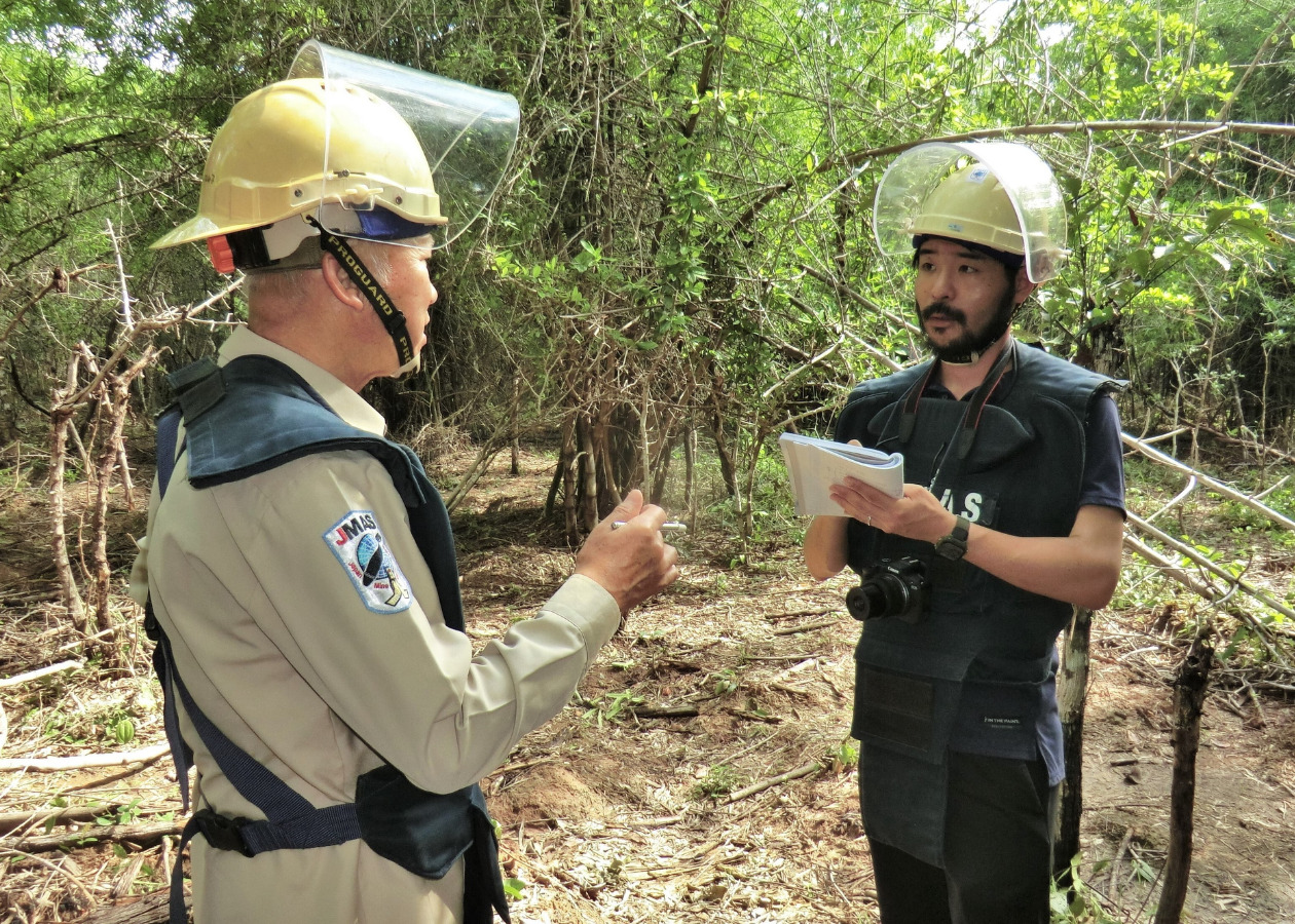 地雷除去の現場で防護服とヘルメットを着用して取材する島袋記者