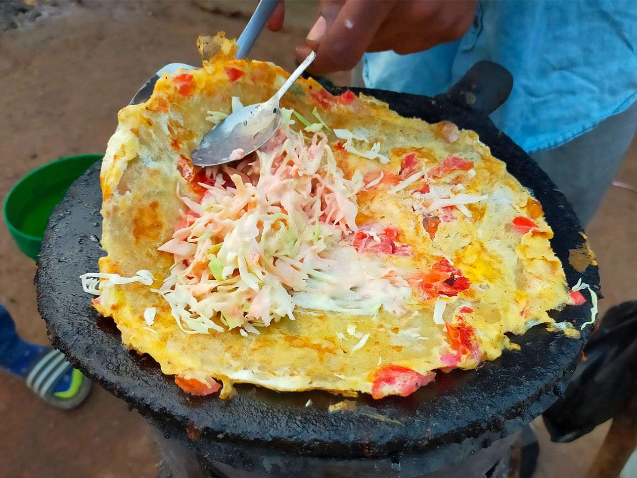 鉄板の上で、黄色い生地にキャベツやトマトがのったお好み焼きに似た食べ物が焼かれている。