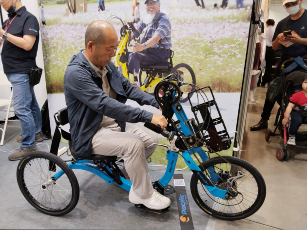 男性が、水色の三輪車に載っている。ハンドル位置には、自転車のようにペダルとチェーンがついている。