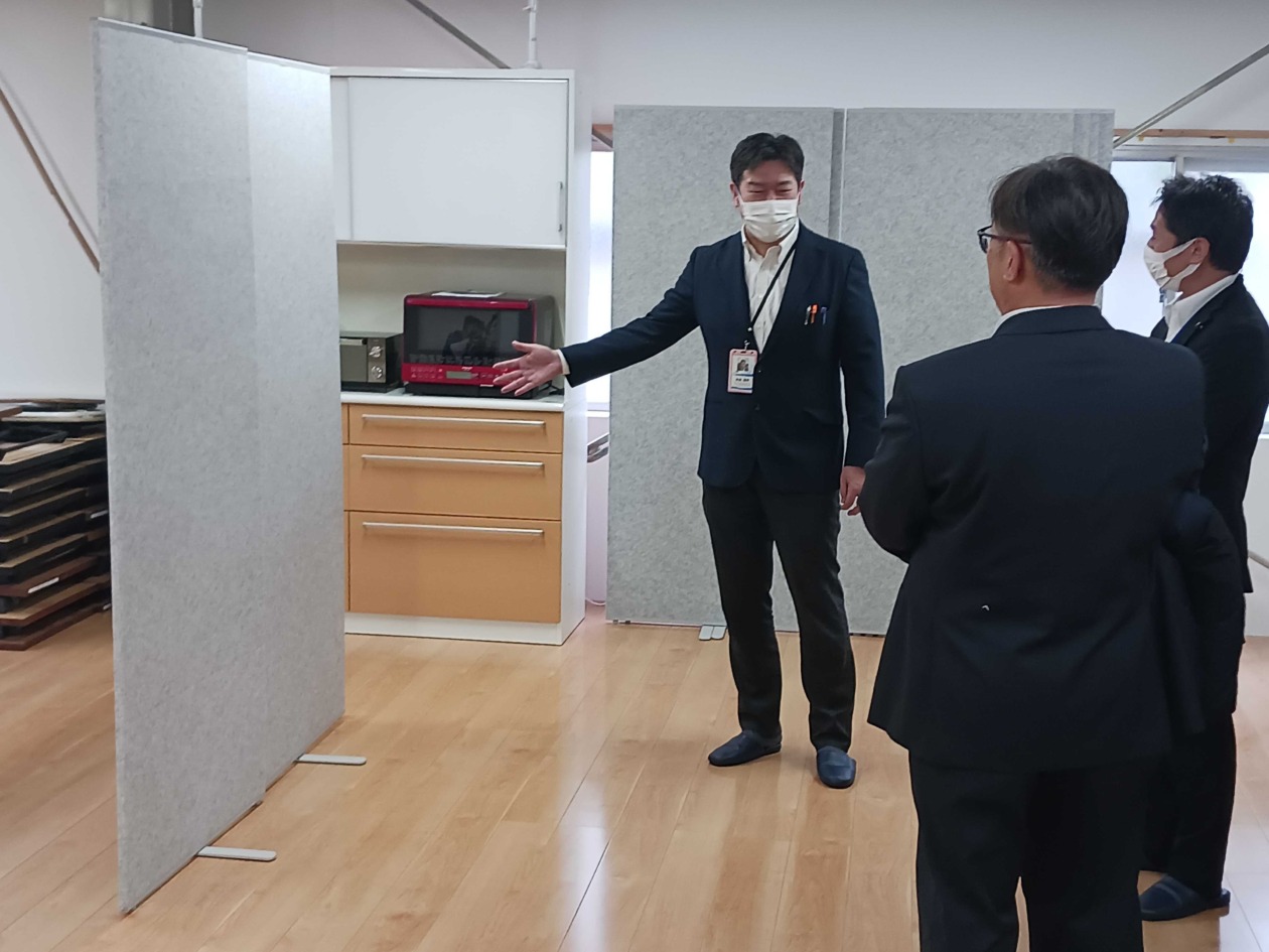 災害時に福祉避難所として活用するために整備された作業所の内部を、関係者が案内している写真です。床はフローリング、動かせる間仕切りが用意されています。
