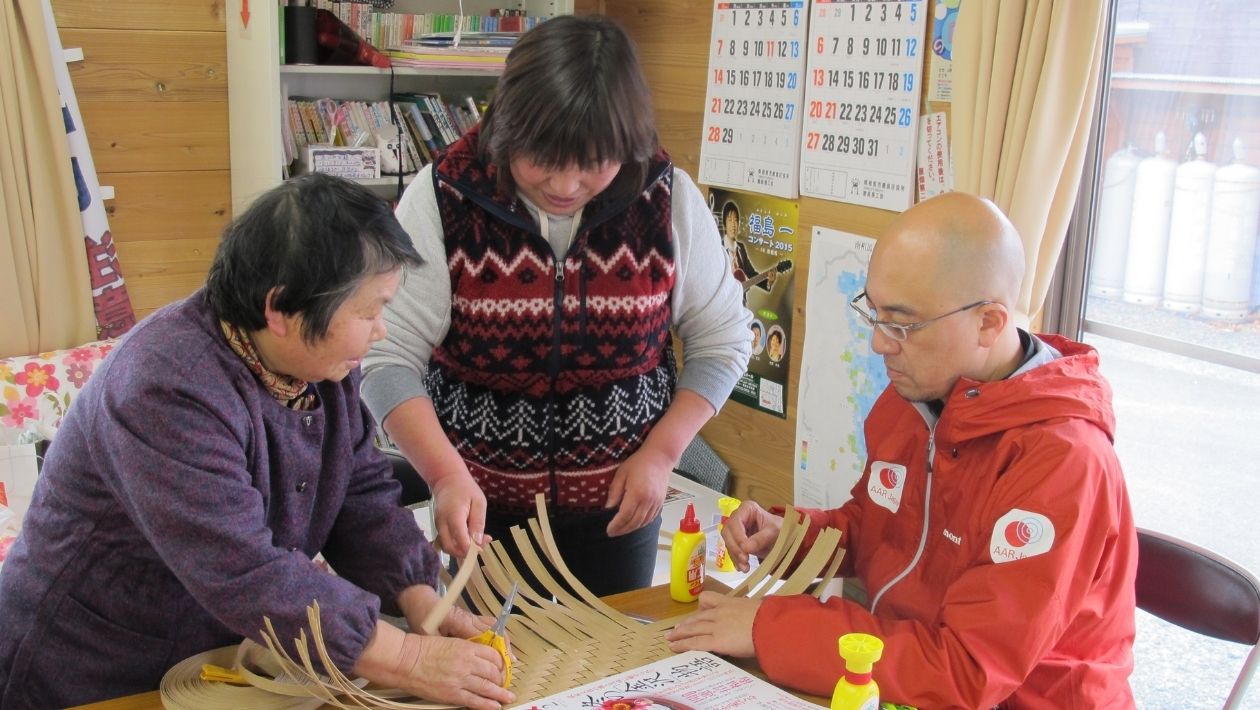 避難所の女性2名とかご編みをする大原真一郎