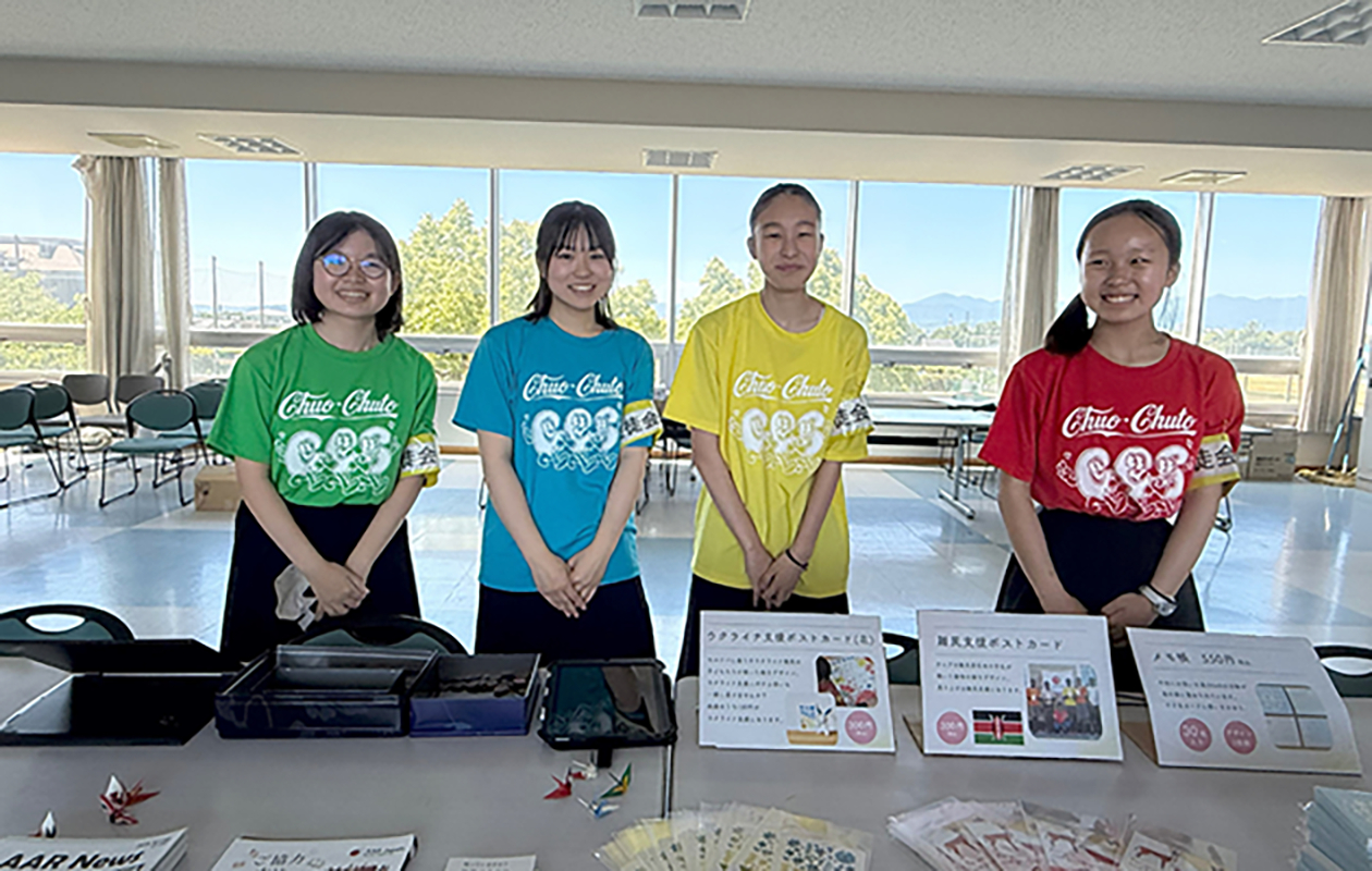 机の後ろに立つ4人の女子生徒。それぞれ違う色のＴシャツを着ている。