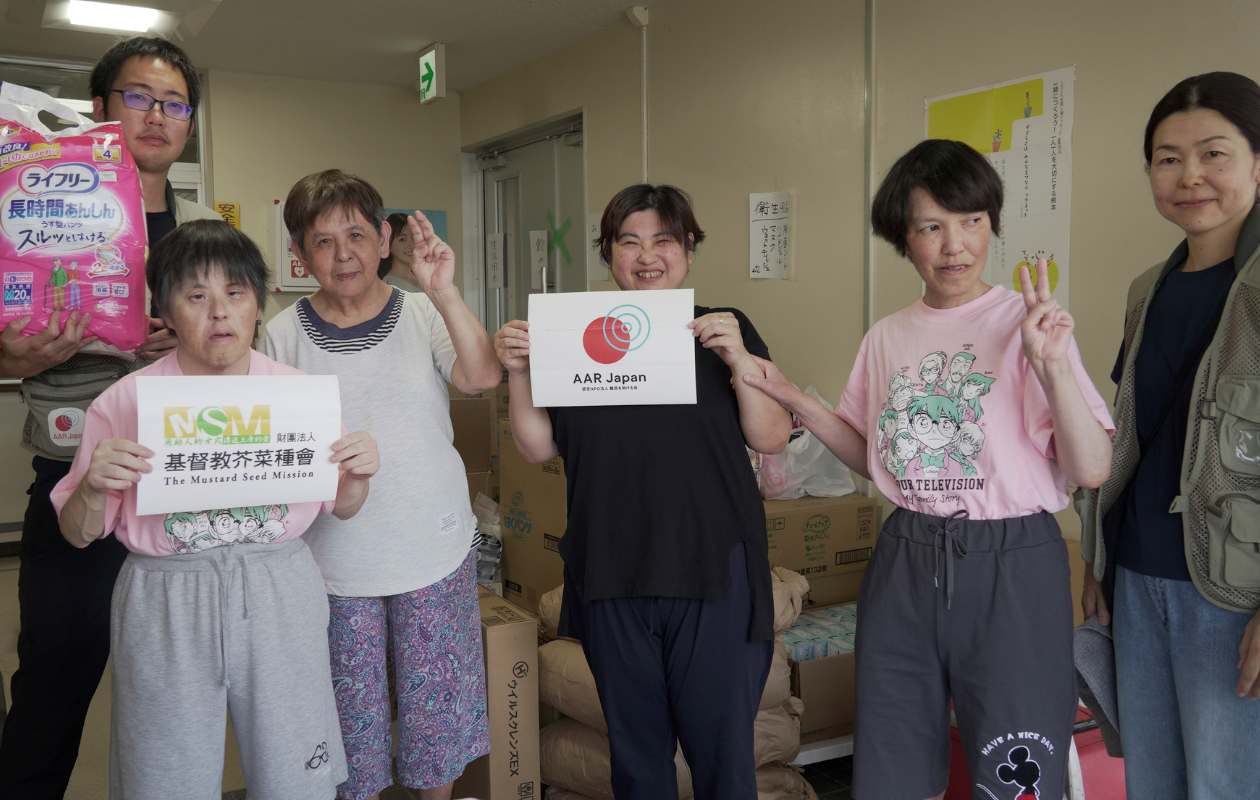 6人の男女が笑顔で並んで立っている。数人は、AARとMSMのロゴが印刷された紙を手に持っている